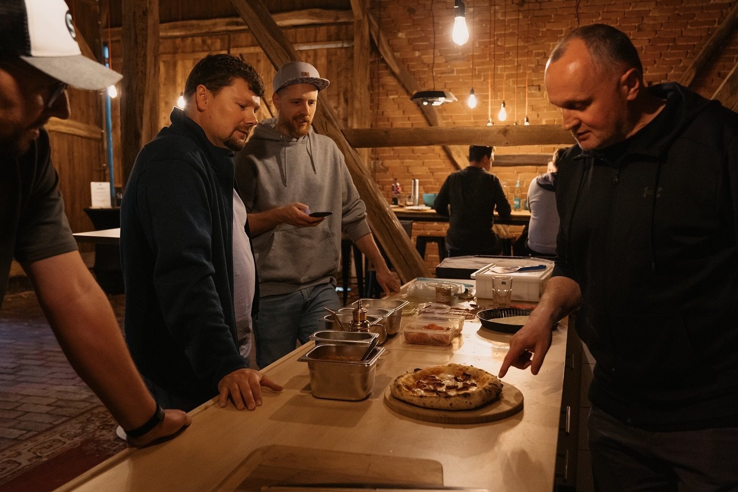 Eine Pizza liegt auf der Arbeitsplatte. Ein Teilnehmer erläutert etwas, während die anderen zusehen.