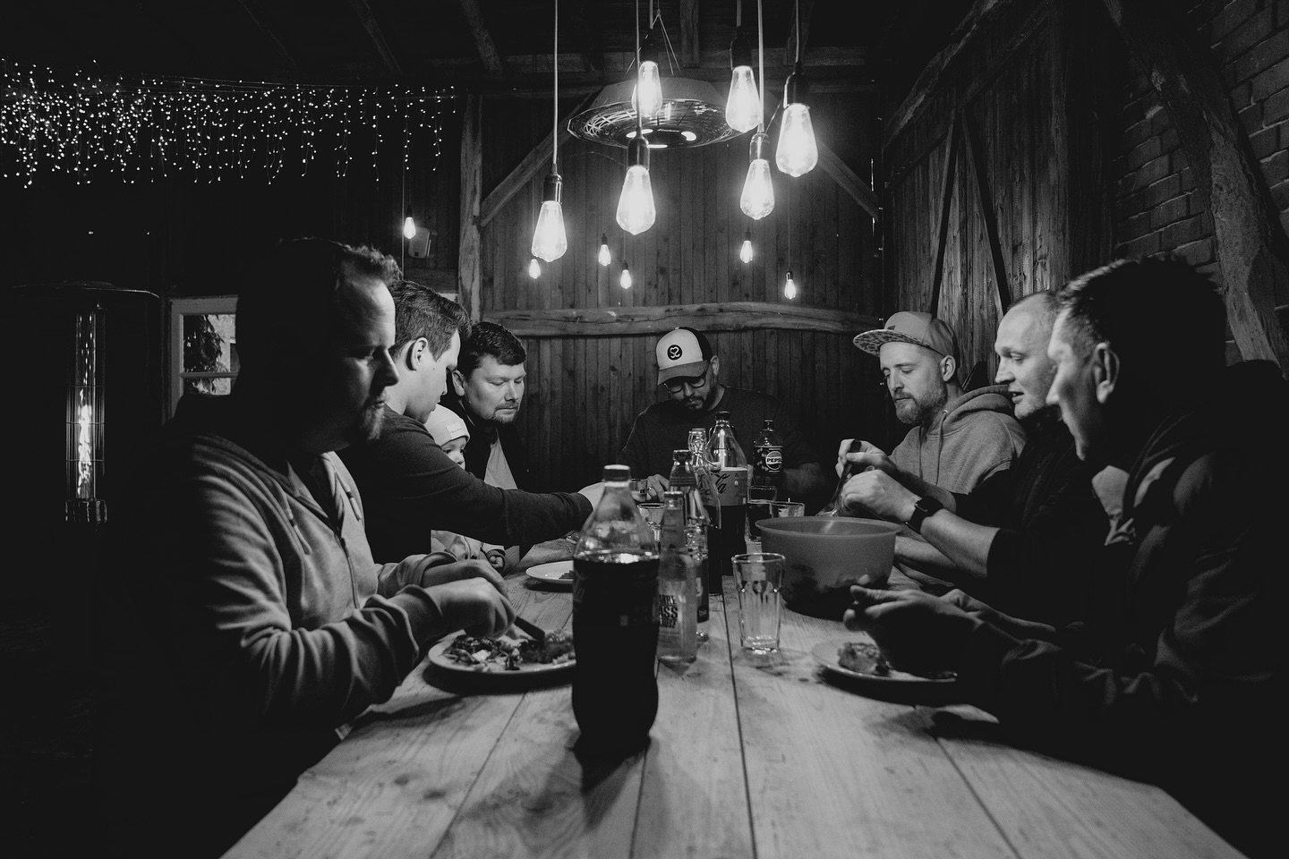 Schwarzweißaufnahme einer Gruppe, die an einer langen Holztafel gemeinsam Pizza isst.